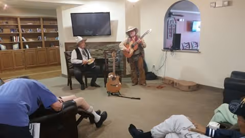 Two musicians performing in a senior living facility common area. One musician is seated playing a percussion instrument, and the other is standing playing a guitar. Several seniors are seated and watching the performance. The room has a TV mounted above a fireplace, bookshelves, and comfortable seating.