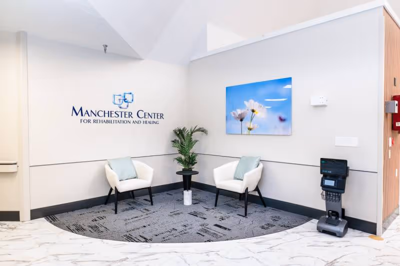 A small waiting or seating area inside Manchester Center for Rehabilitation and Healing featuring two white chairs with light blue cushions, a small round black table with a potted plant, a wall-mounted picture of white flowers against a blue sky, and a device on the right side near the wall.