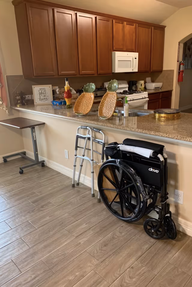 Interior view of a kitchen area with wooden cabinets, a microwave, stove, and granite countertop. On the counter are two pineapple-shaped decorations and various kitchen items. In front of the counter, there is a walker and a folded wheelchair. The floor has wood-like tiles, and there is a small rolling table to the left.