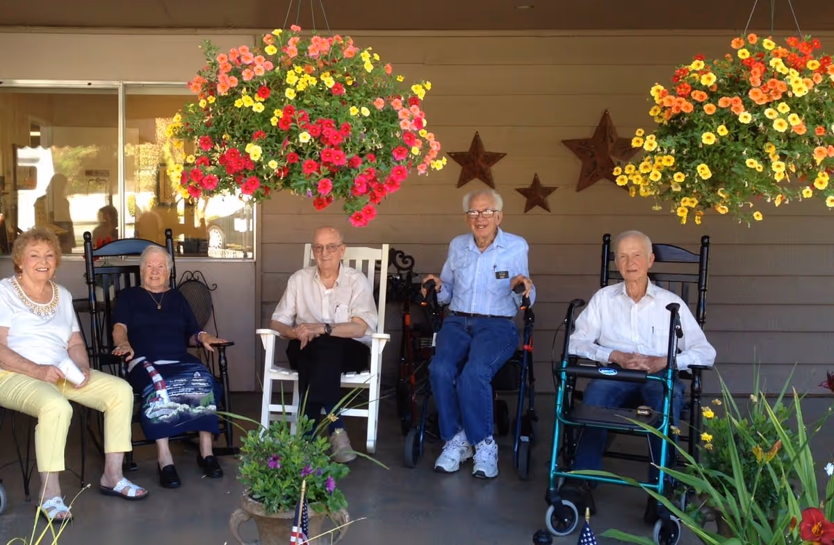 Five elderly individuals sitting on chairs and walkers on a covered porch decorated with hanging flower baskets and star wall decorations, enjoying a sunny day at a retirement facility.