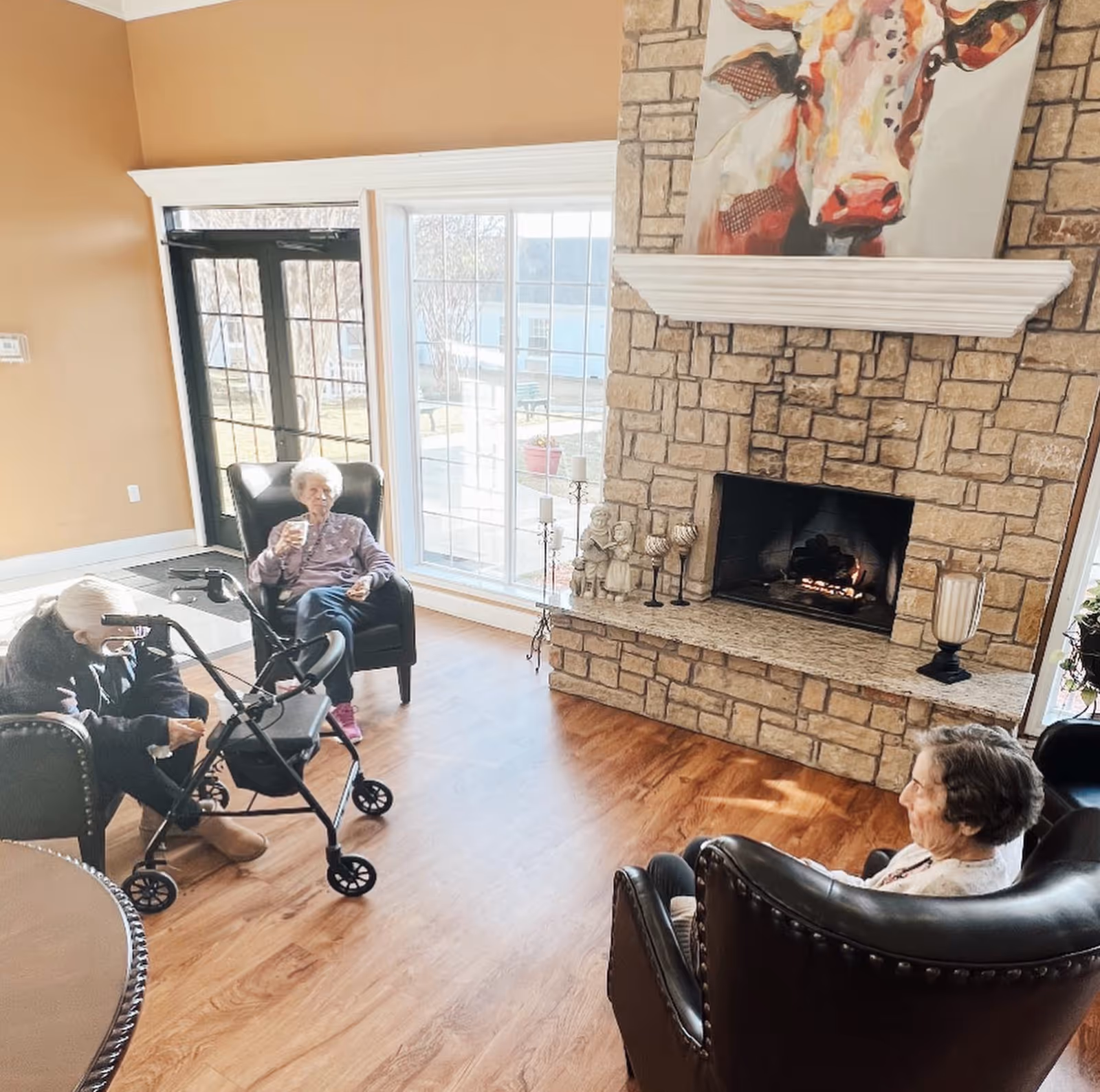 Three elderly women sitting in a cozy living room with wooden floors and a stone fireplace. One woman is seated in a black armchair near a large window with sunlight streaming in, holding a drink. Another woman is seated with a walker nearby, and the third woman is sitting in a leather armchair facing the fireplace. Above the fireplace is a colorful painting of a cow.