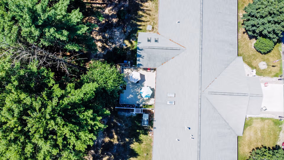 Aerial view of a long gray-roofed building next to dense trees with a small patio area visible.