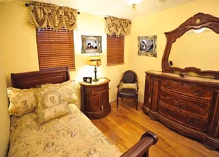 A cozy bedroom with a wooden bed featuring patterned bedding and multiple pillows. The room has two windows with wooden blinds and decorative valances. Between the windows is a small round wooden nightstand with a lamp and a framed picture above it. There is a wooden dresser with an ornate mirror on the right side and a single chair with a cushion next to the nightstand. The walls are painted yellow and the floor is hardwood.