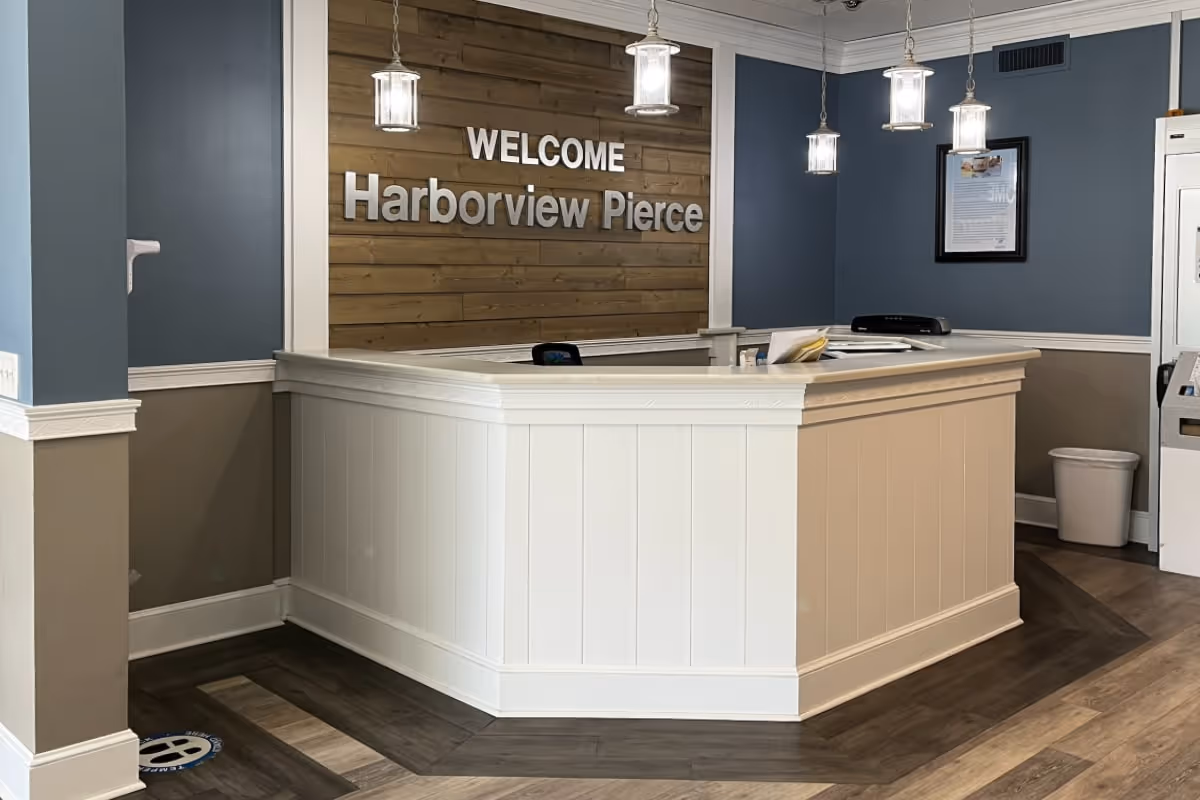 Reception area with a white counter and wooden panel wall behind it displaying the text 'WELCOME Harborview Pierce'. The walls are painted blue and beige, with three pendant lights hanging from the ceiling. There is a framed document on the wall and a trash bin in the corner.