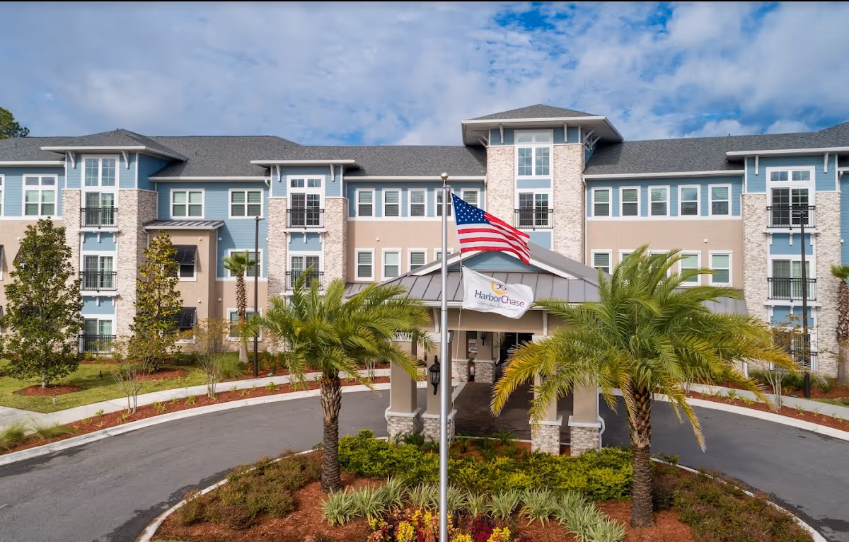 Front exterior view of HarborChase of Mandarin, a multi-story senior living facility with a circular driveway, landscaped garden with palm trees, and an American flag flying in front of the entrance.