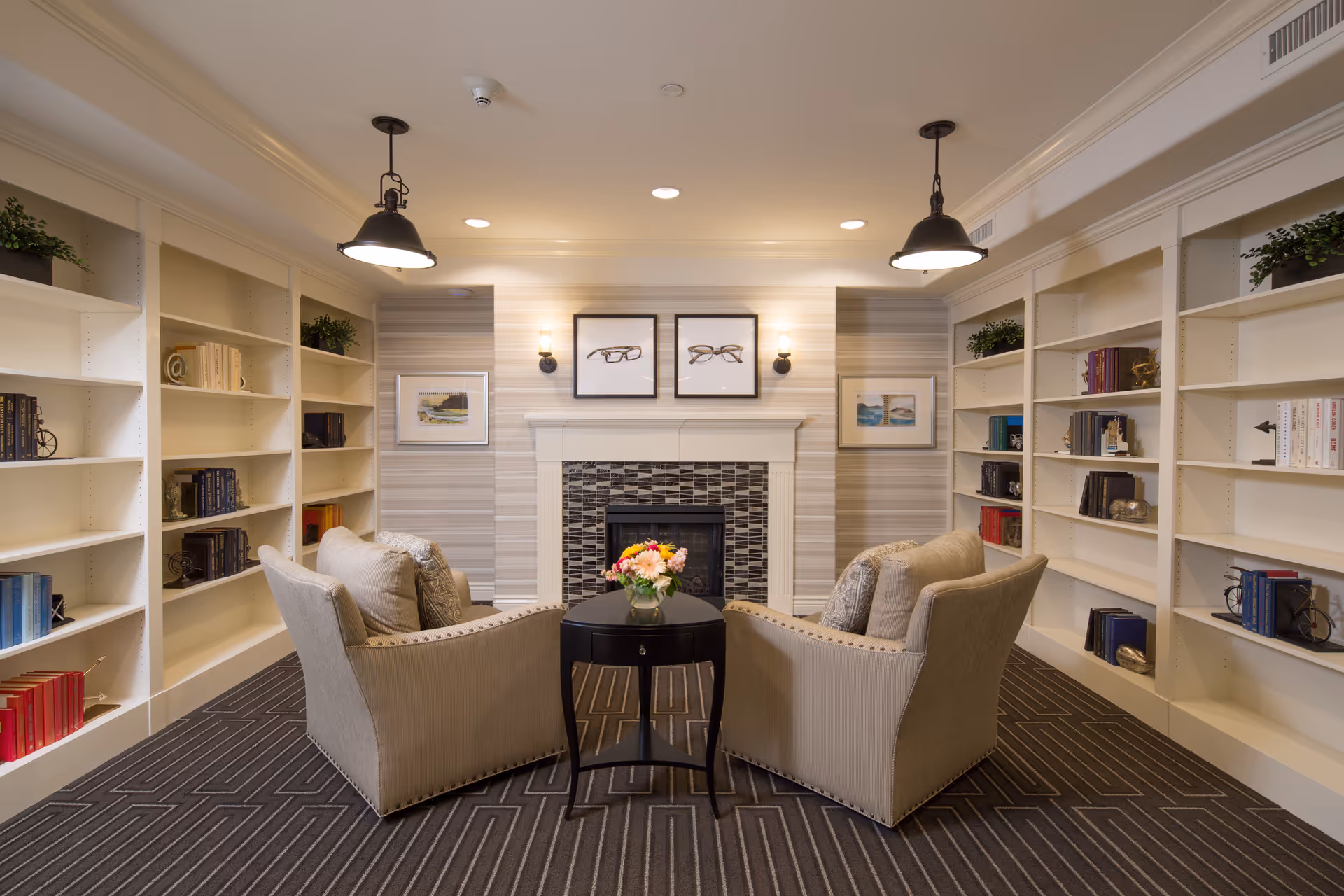 A cozy sitting area in a senior living facility with two beige armchairs facing a small round black table with a vase of flowers. The room features built-in white bookshelves filled with books and decorative items on both sides, a fireplace with a tiled surround, and framed artwork above it. The ceiling has recessed lighting and two hanging pendant lamps.