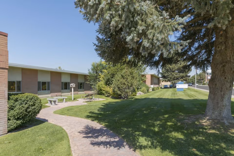 Outdoor view of Gateway Transitional Care Center showing a paved walkway, green lawn, trees, bushes, and a picnic table with benches near the building under a clear blue sky.