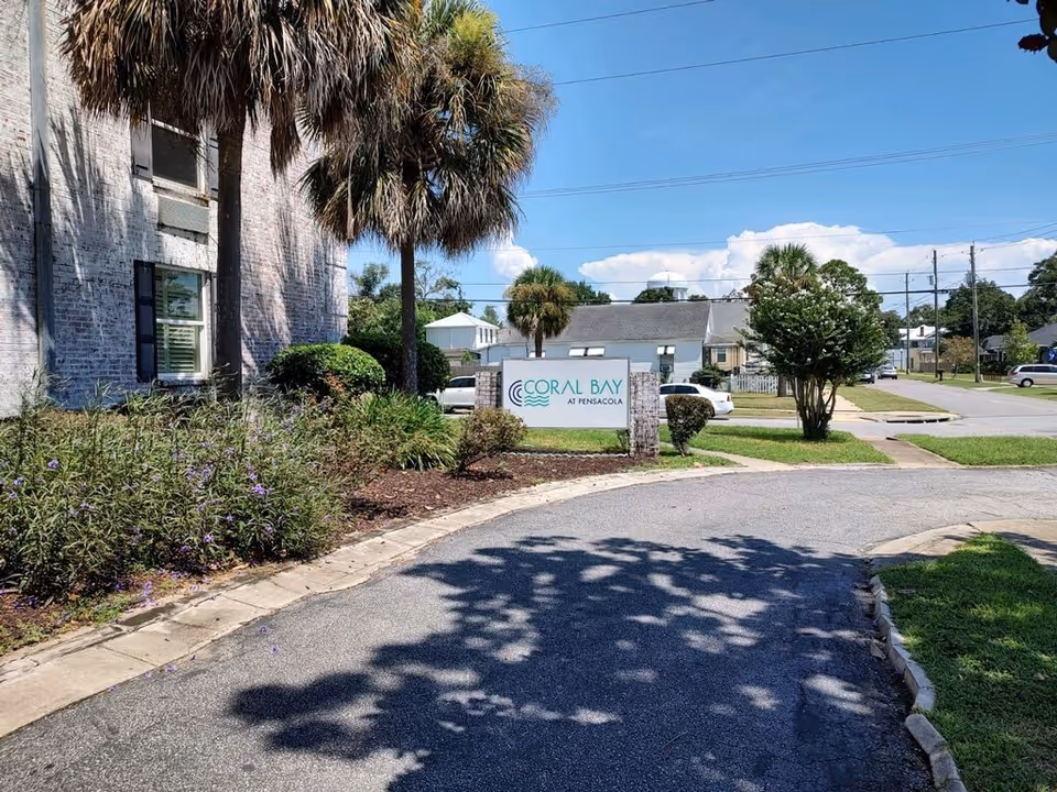 Outdoor view of the entrance area to Coral Bay at Pensacola facility, showing a curved driveway, landscaped bushes and palm trees, a white brick building on the left, and a sign with the facility name. Residential houses and a clear blue sky are visible in the background.