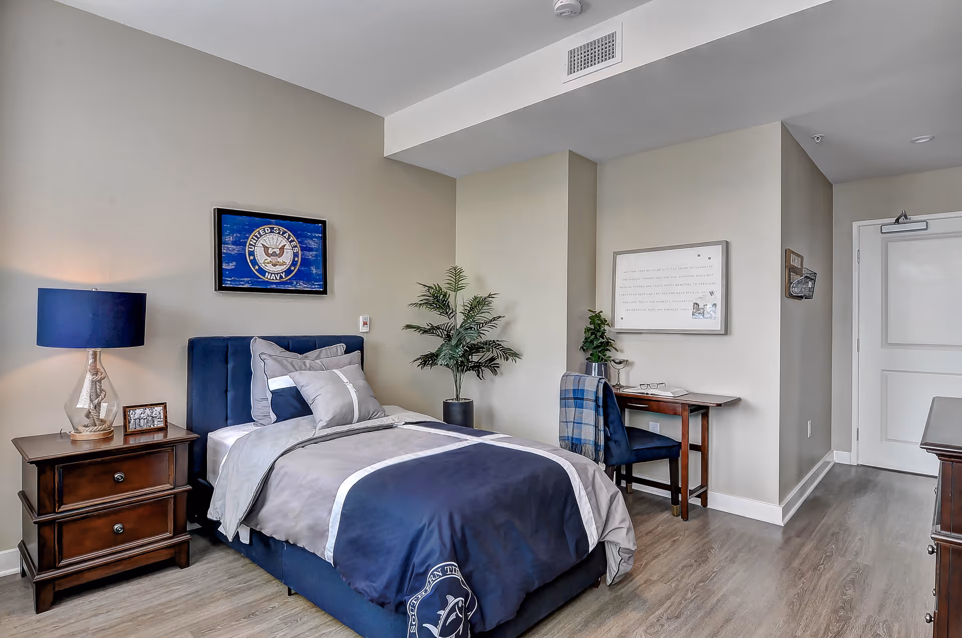 A neatly arranged bedroom with a single bed featuring navy blue and gray bedding. There is a wooden nightstand with a lamp and a framed photo beside the bed. A framed United States Navy emblem hangs on the wall above the bed. A small desk with a chair and a plant is positioned against the wall near the door. The room has light-colored walls and wood flooring.