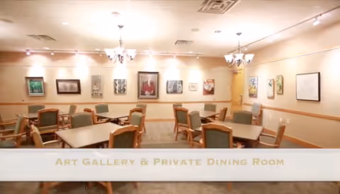 A well-lit room with multiple square tables and green cushioned chairs arranged neatly. The walls are adorned with various framed artworks, creating an art gallery ambiance combined with a private dining room setting. Ceiling lights and chandeliers illuminate the space.