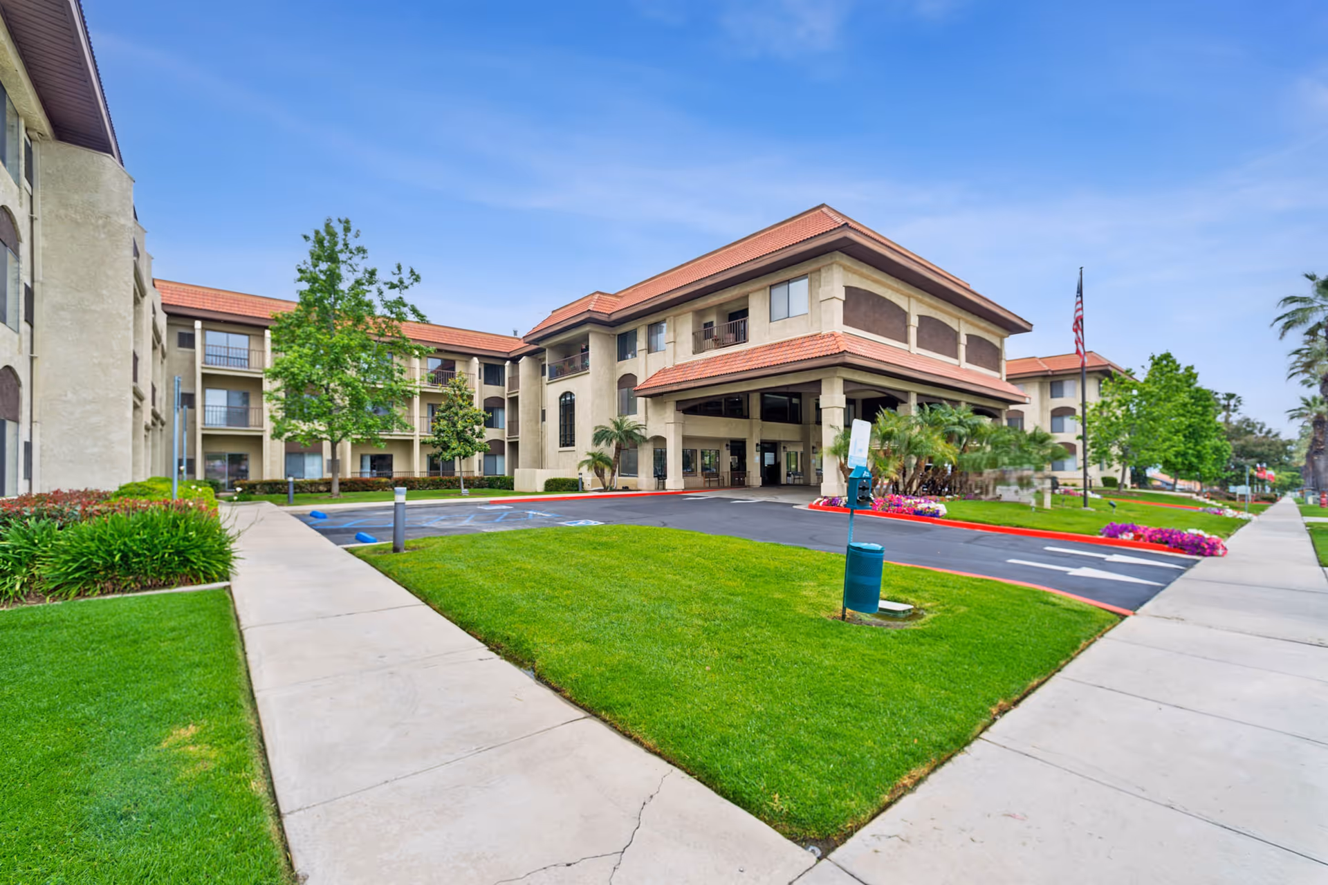 Front exterior of a multi-story senior living facility with a covered entrance, landscaped lawn, and flagpole.