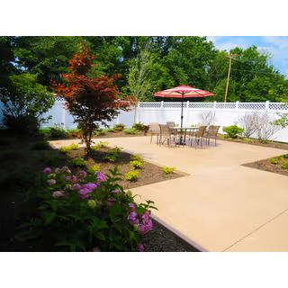 Outdoor patio area with a concrete surface, a table with six chairs, and a red umbrella. The patio is surrounded by landscaped garden beds with various plants and flowers, and a white fence encloses the area. Trees and greenery are visible in the background under a blue sky.