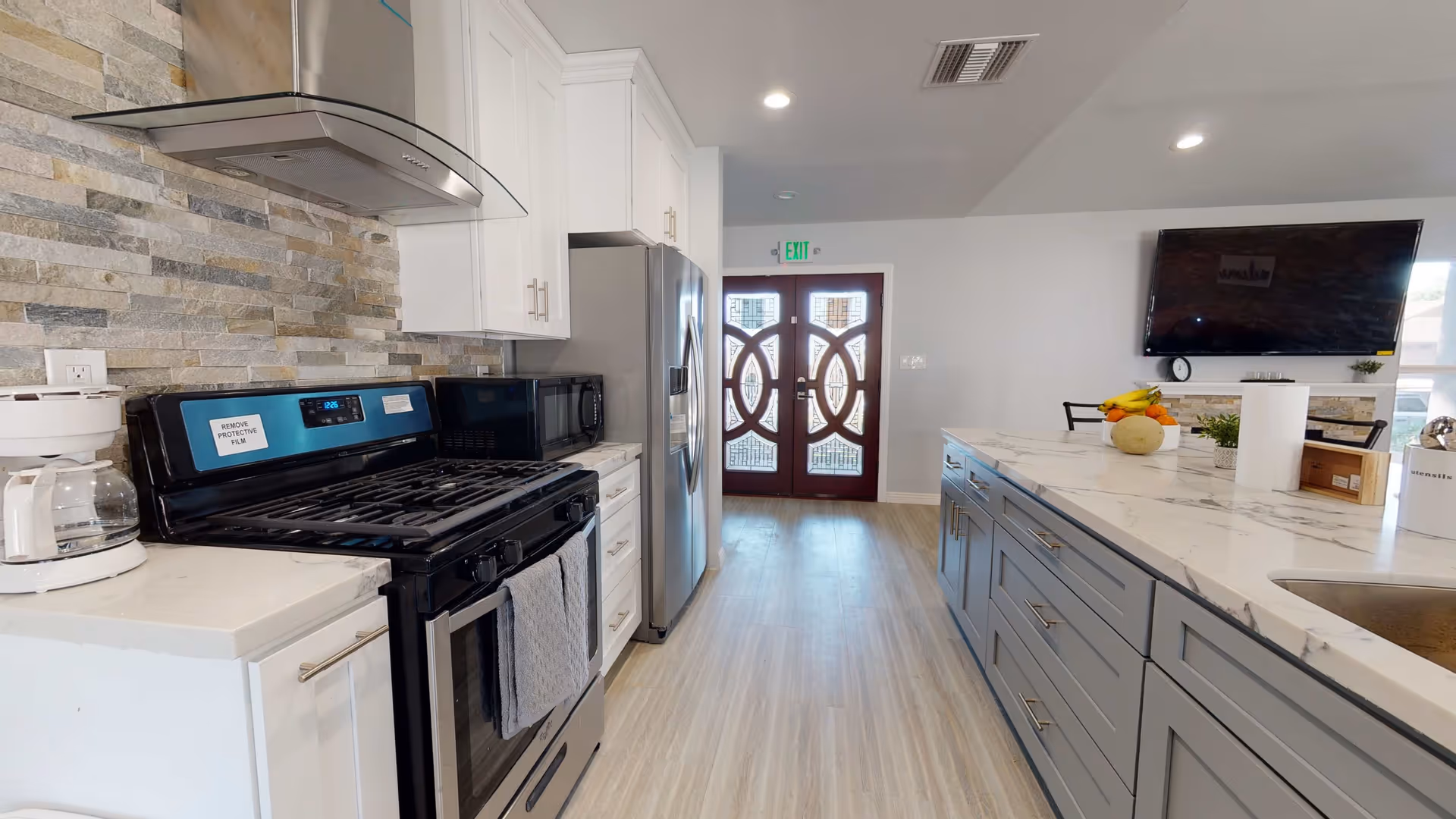 Modern kitchen with white cabinets, a stainless steel refrigerator, a black stove with a range hood, a microwave, and a coffee maker. There is a large island with gray drawers and a marble countertop, decorated with fruit and paper towels. Double doors with decorative glass panels are visible at the far end of the room.