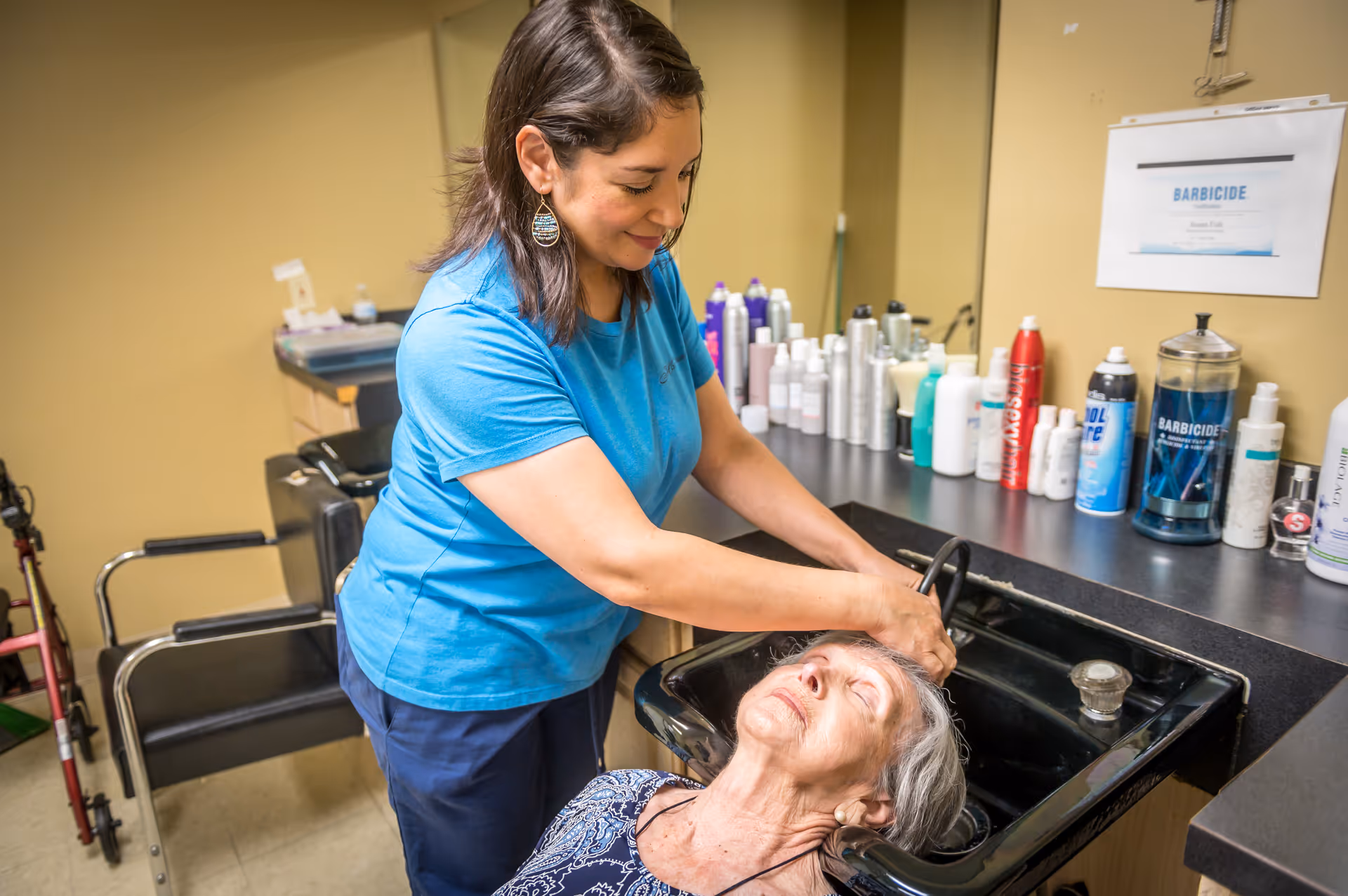 A caregiver washing the hair of an elderly woman who is reclining with her head in a black salon sink. Various hair care products are lined up on the counter behind them.
