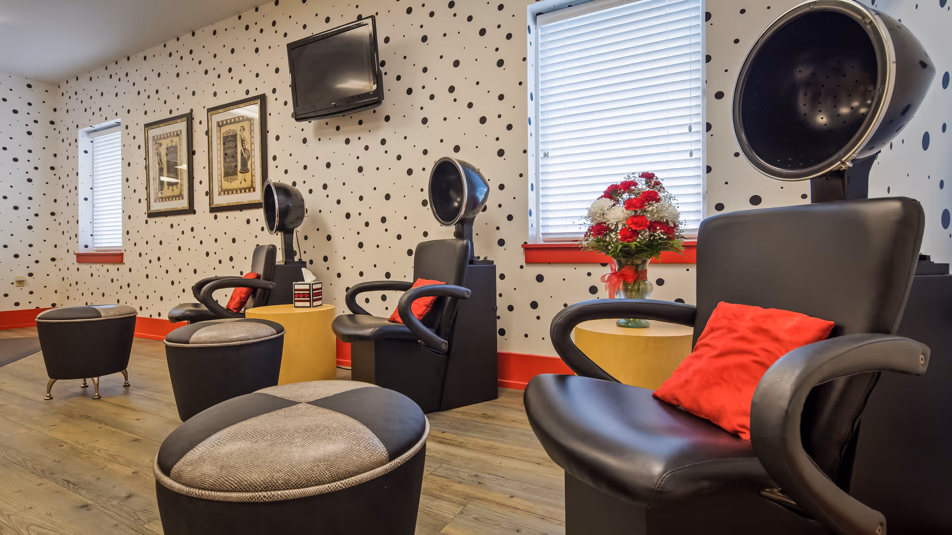 Interior of a salon area with black hair drying chairs, each with a red pillow, small round ottomans, a wooden floor, polka dot patterned walls, two windows with blinds, framed pictures on the wall, a small round table with a vase of red and white flowers, and a wall-mounted TV.
