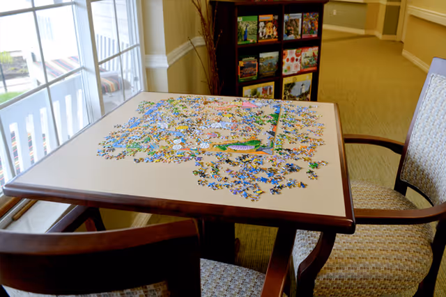 A table with a partially completed jigsaw puzzle on it, surrounded by two cushioned chairs. The table is near a window with a view outside, and a bookshelf with various books is visible in the background.