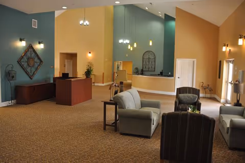 A spacious senior living facility lobby with beige and teal walls, carpeted floor, a reception desk, and several seating areas with armchairs and small tables. The ceiling is vaulted with hanging pendant lights, and decorative wall art is visible.
