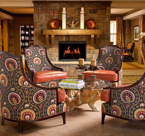 A cozy living room area with four patterned armchairs arranged around a glass coffee table with a natural wood base. The background features a stone fireplace with a fire burning, decorated with candles and ornamental plates. Books are stacked on the coffee table, and the room has warm lighting and earth-toned decor.