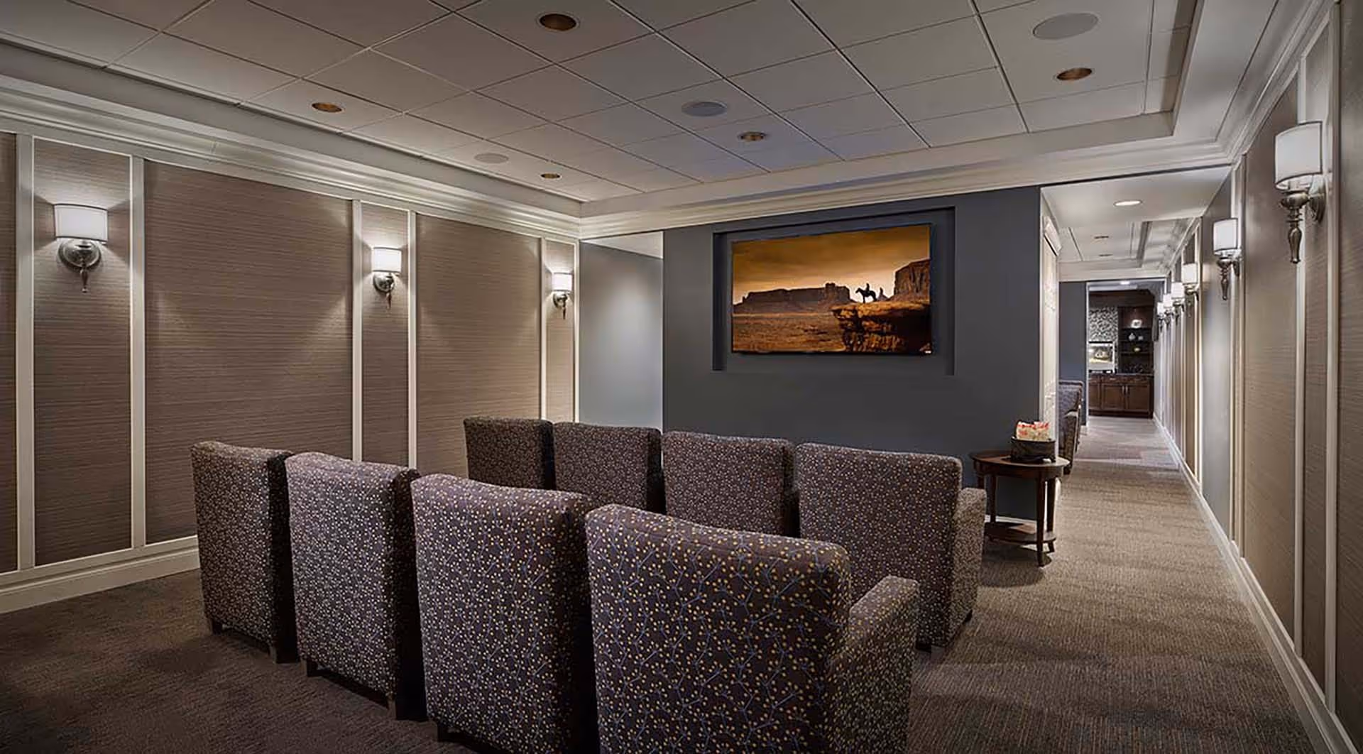 A small theater room with two rows of patterned armchairs facing a wall-mounted flat screen TV displaying a western scene. The room has soft wall lighting, a carpeted floor, and a hallway leading to another area with cabinetry and a coffee machine.