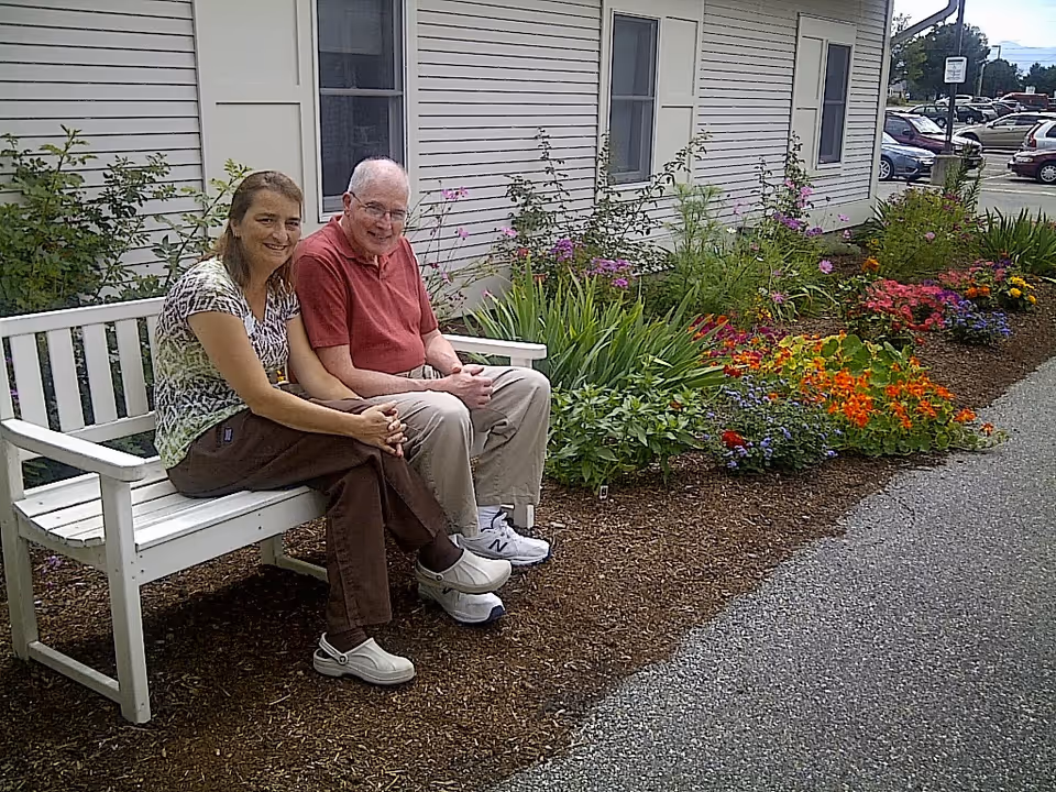 Two people sitting on a white bench beside a colorful flowerbed outside a light-colored building.