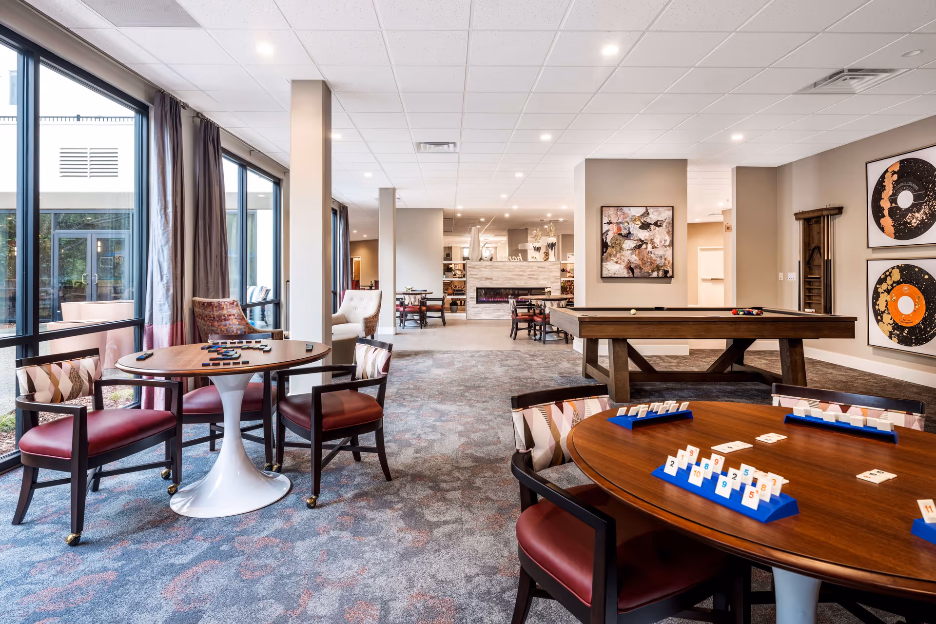 A spacious and well-lit common area in a senior living facility featuring multiple round tables with chairs arranged for playing games, a pool table, large windows with curtains allowing natural light, modern artwork on the walls, and a cozy fireplace in the background.