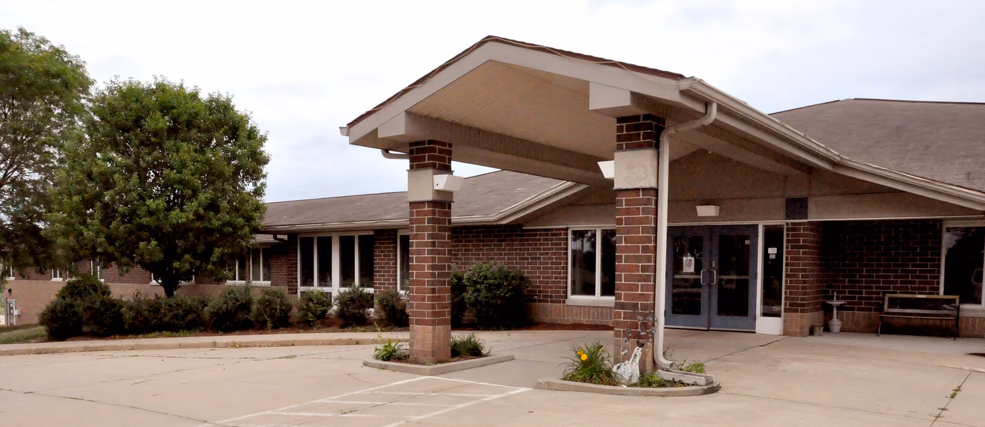 Entrance of a single-story brick building with a covered driveway supported by two brick pillars. There are bushes and a tree to the left of the entrance, and a bench and a small birdbath near the door. The sky is overcast.