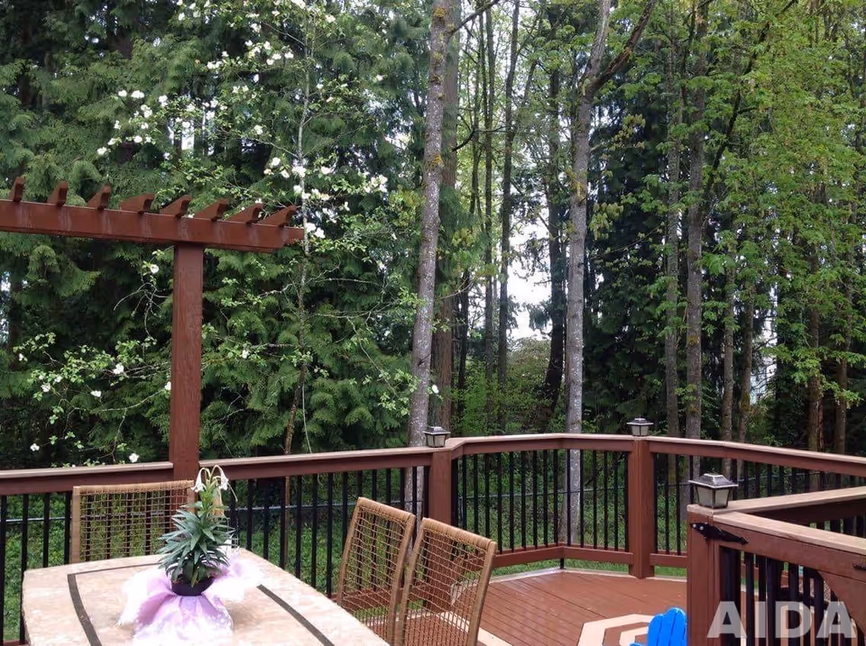 Outdoor wooden deck area with a table and chairs, surrounded by a railing and trees in the background. A pergola structure is visible on the left side, and there is a potted plant with a purple cloth on the table.