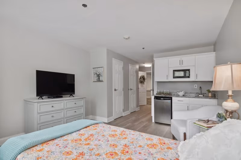 Interior view of a senior living facility room featuring a bed with a floral bedspread and a light blue blanket, a white dresser with a flat-screen TV on top, a small kitchenette with white cabinets, a microwave, a mini fridge, and a sink. There is a white armchair next to a side table with a lamp and some books. The walls are painted light gray and the floor is wood.