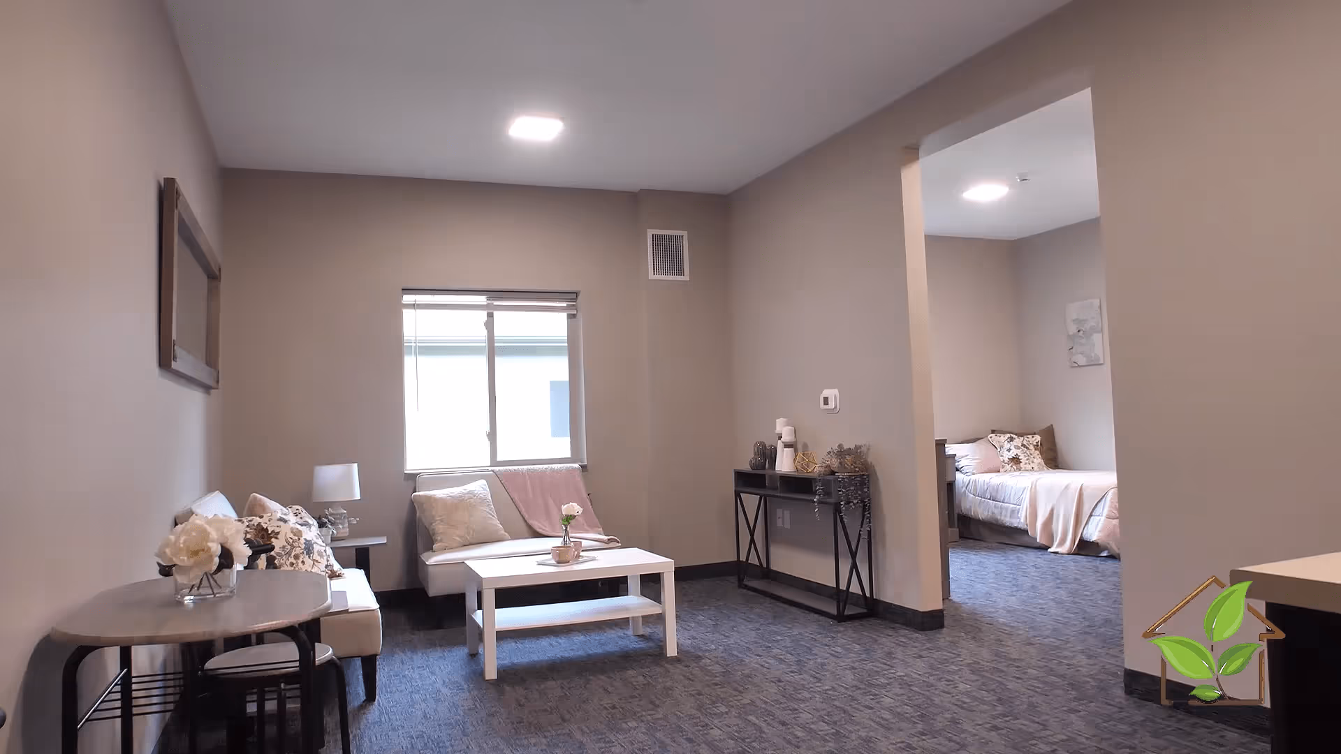 A cozy living room area with a small white couch, a white coffee table with a flower vase, and a side table with a lamp. The room has beige walls and a window with blinds. To the right, there is an open doorway leading to a bedroom with a bed, pillows, and a blanket. The floor is carpeted in a blue-gray color.