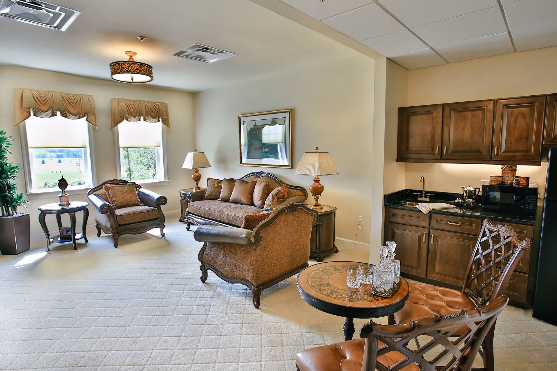 A cozy living area with a sofa, two armchairs, a small round table with two chairs, and a kitchenette with wooden cabinets, a sink, a microwave, and a refrigerator. The room has two windows with beige valances, two table lamps, a framed picture on the wall, and a potted plant near the windows.