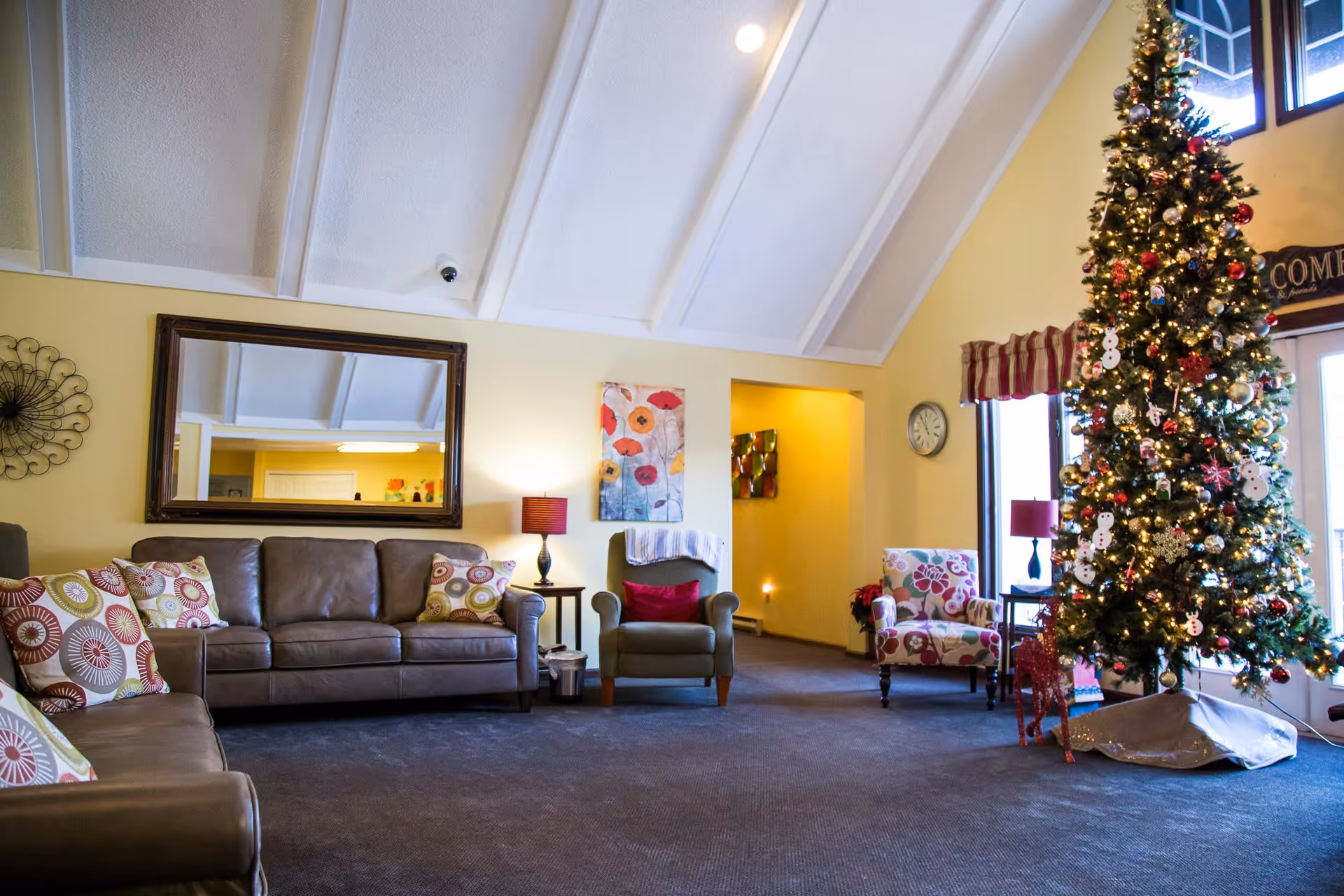 A cozy living room decorated for Christmas with a tall, decorated Christmas tree on the right side. The room features a brown leather sofa with patterned cushions, two armchairs with colorful floral patterns, a large mirror on the wall, a wall clock, and warm yellow walls with a high vaulted ceiling.
