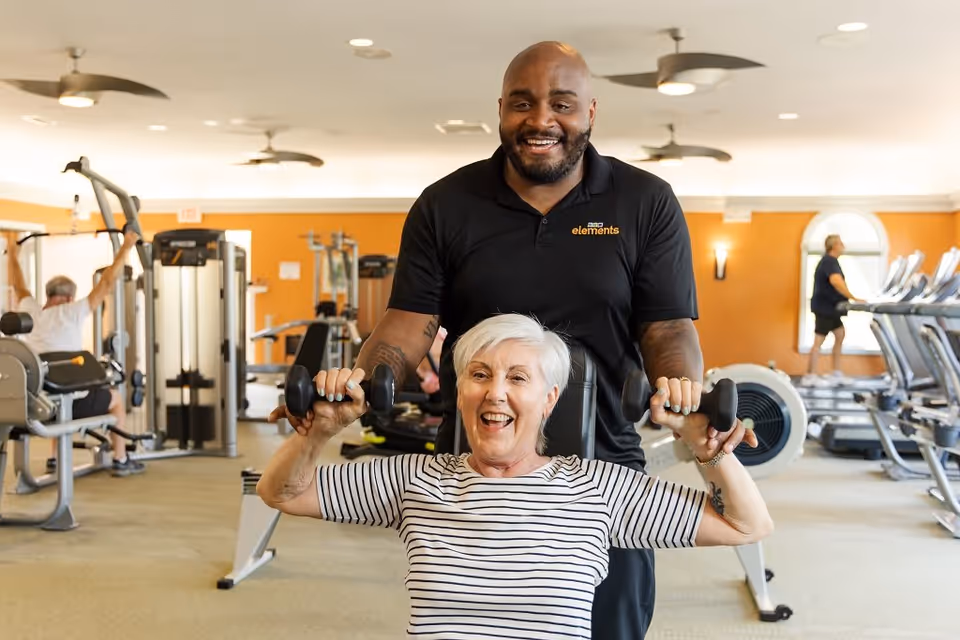 An elderly woman with short white hair and a striped shirt is seated and lifting dumbbells with the assistance of a smiling male trainer wearing a black polo shirt. They are in a well-lit gym with exercise machines and treadmills in the background, where other people are also working out.