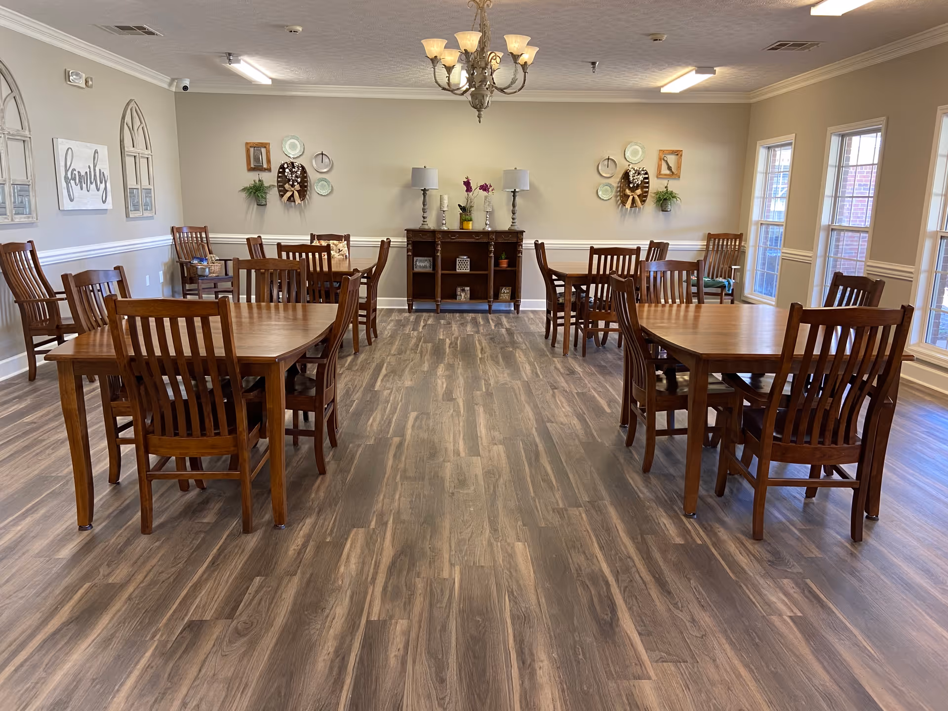 A dining room with several wooden tables and chairs arranged neatly on a wood-patterned floor. The room has beige walls decorated with wall art, plants, and two table lamps on a wooden sideboard against the far wall. Large windows on the right side allow natural light to fill the space, and a chandelier hangs from the ceiling.