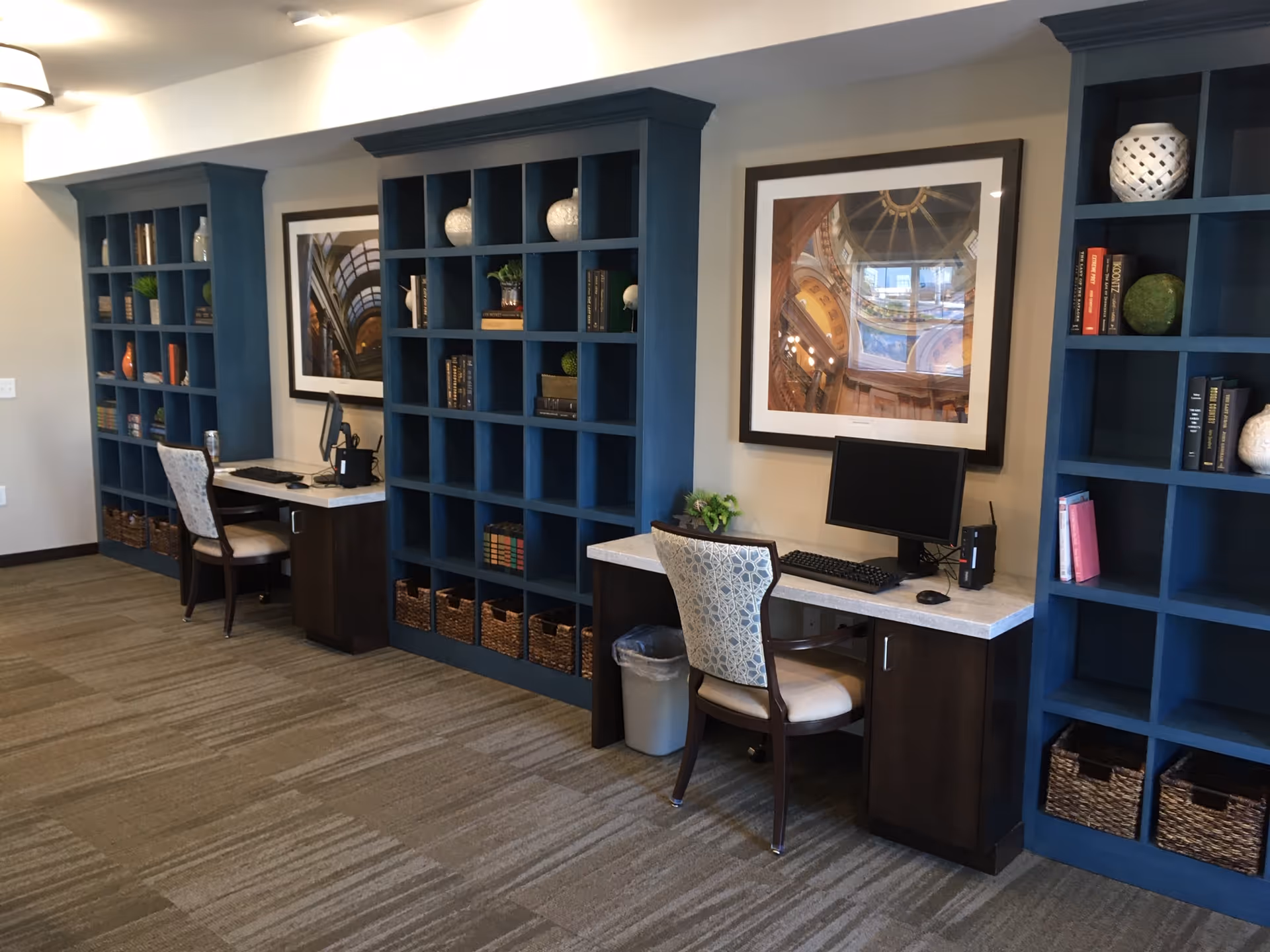 Interior view of a senior living facility workspace area with two desks, each equipped with a computer monitor, keyboard, and mouse. The desks are positioned against a wall with large blue built-in bookshelves filled with books, decorative items, and baskets. Above each desk hangs a framed photograph of architectural details. The floor is carpeted, and there are upholstered chairs at each desk.