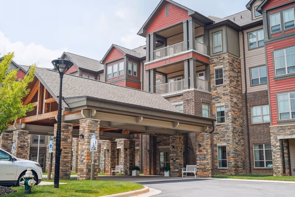Exterior view of a multi-story senior living facility with stone and red siding accents. The building features covered entrance with stone pillars, balconies, and a parking area with a white vehicle and a fire hydrant visible.