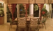 Dining area with a rectangular glass-top table surrounded by six metal chairs with cushions. The room has large windows with red and beige striped curtains, and green plants are visible outside the windows.