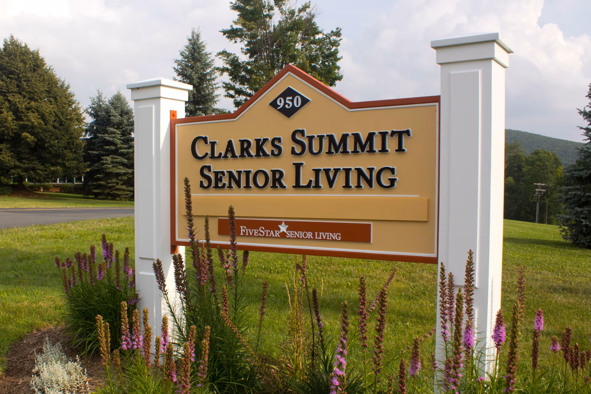 Large freestanding sign reading 'Clarks Summit Senior Living' surrounded by flowers and greenery in front of trees and a lawn.