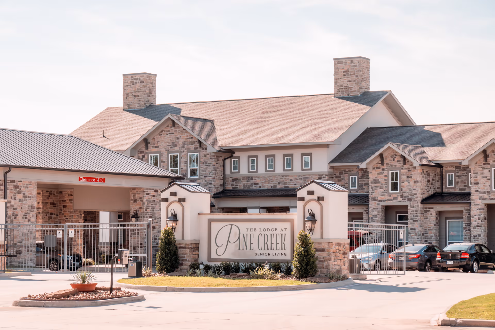 Exterior view of The Lodge at Pine Creek senior living facility showing a large stone building with multiple windows, two stone chimneys, and a gated entrance with a sign displaying the facility's name. Several cars are parked near the entrance.