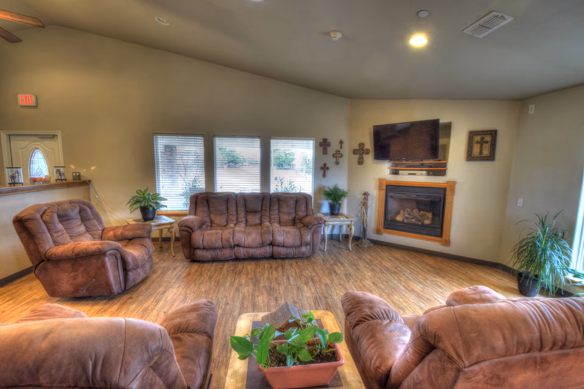 A cozy living room with brown cushioned sofas and armchairs arranged around a wooden coffee table with a potted plant. The room features wood flooring, a wall-mounted TV above a fireplace, several decorative crosses on the wall, and multiple windows letting in natural light.