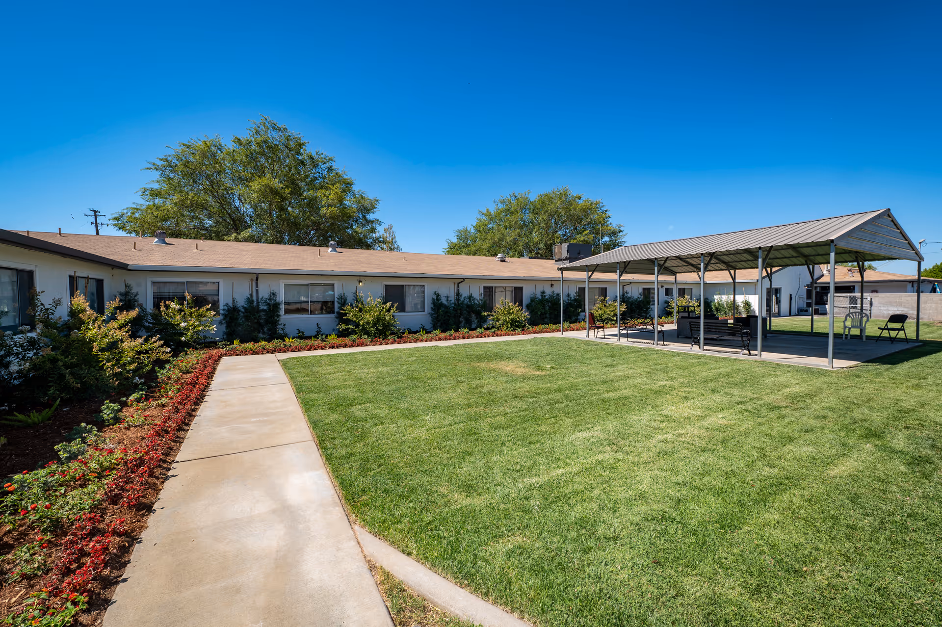 Outdoor view of a senior living facility courtyard with a well-maintained lawn, a concrete walkway, flower beds along the building, and a covered seating area with chairs and benches under a metal roof. The building is single-story with multiple windows and surrounded by trees under a clear blue sky.