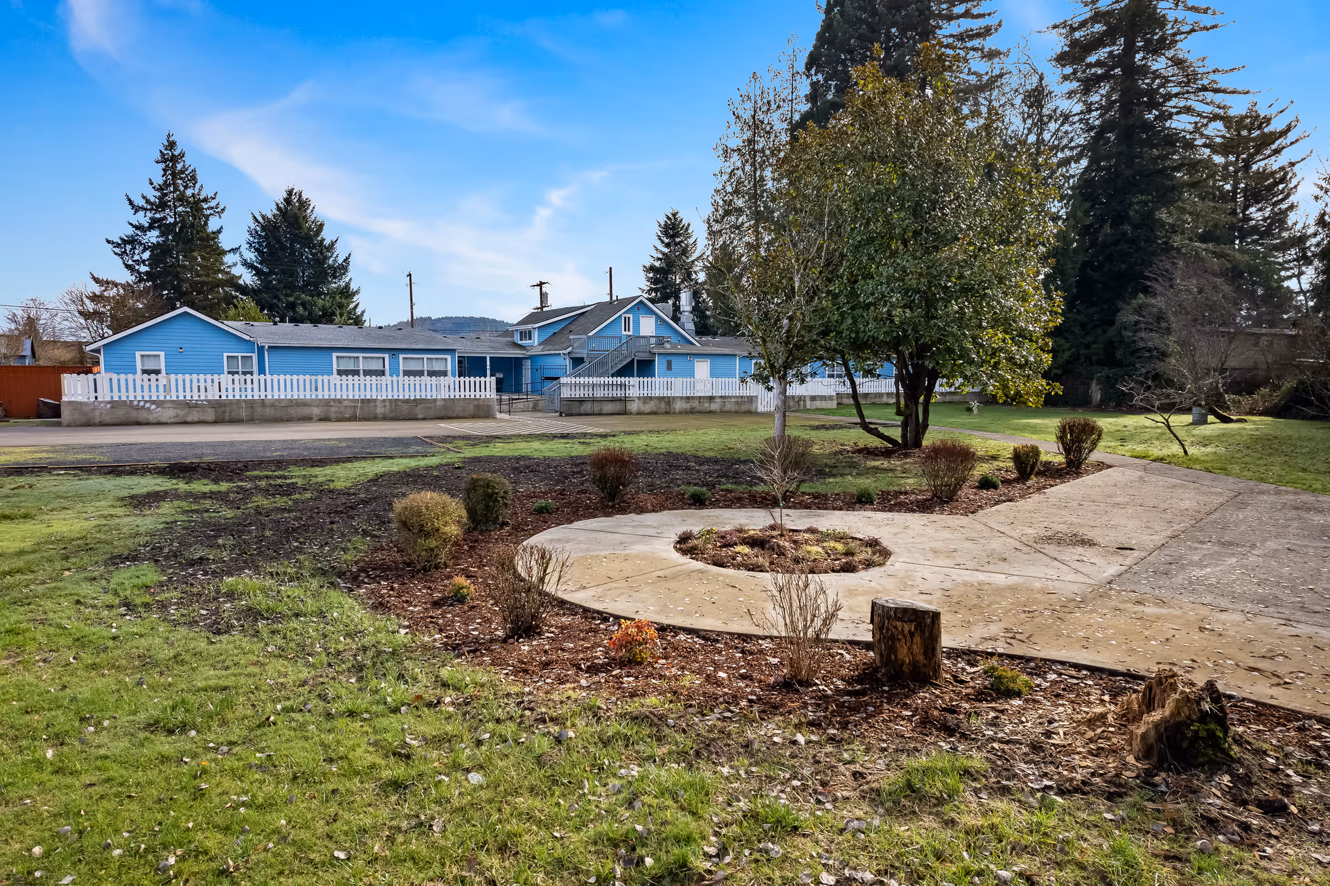 Outdoor view of Wellspring Centralia facility showing a blue building with white fencing, surrounded by green grass, trees, and a circular concrete pathway with small bushes and plants.