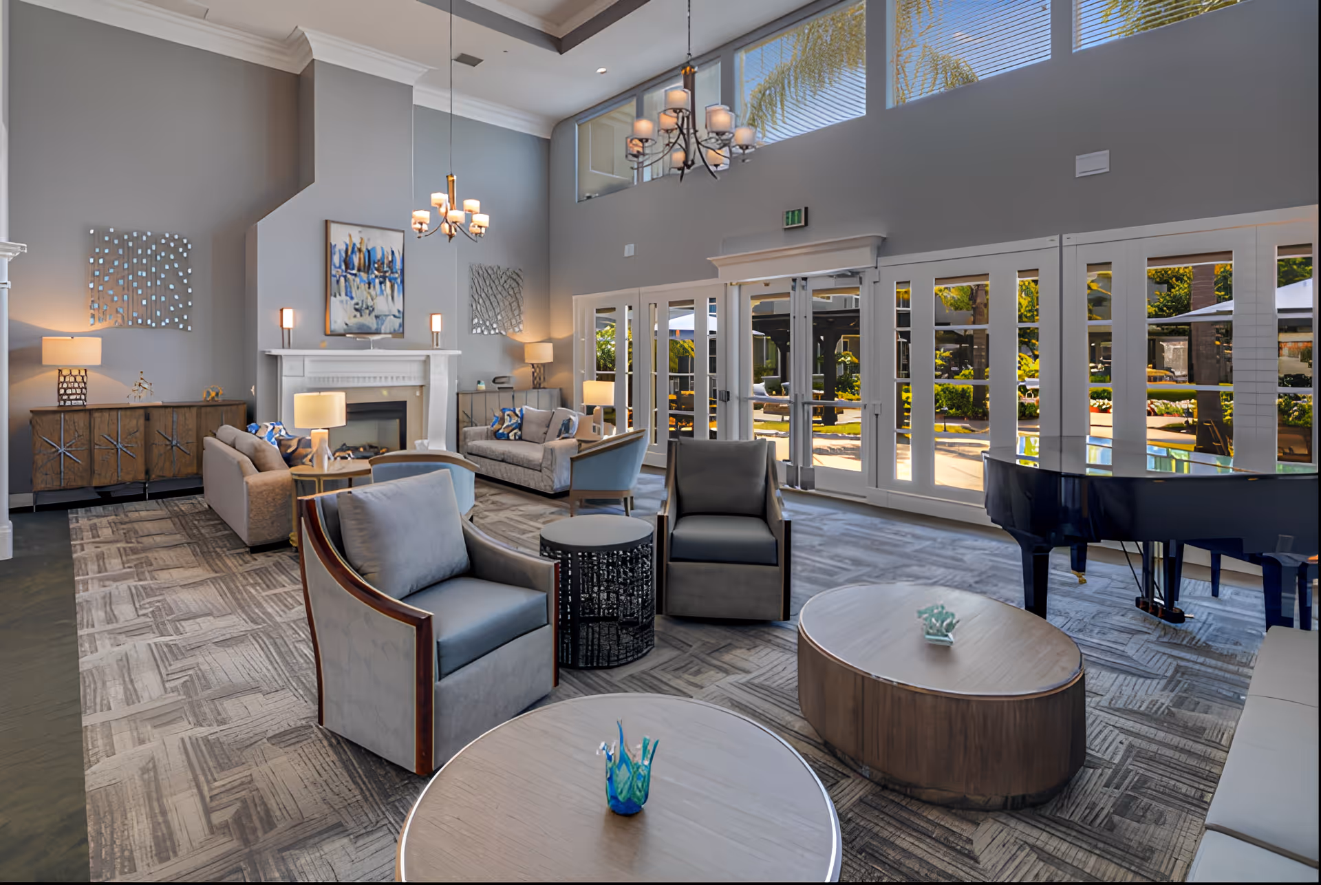 A spacious and well-lit living room area with modern furniture including armchairs, sofas, and coffee tables. The room features a fireplace with artwork above it, decorative wall pieces, and multiple lamps. Large windows and glass doors allow natural light to enter and provide a view of an outdoor garden area. A grand piano is positioned near the windows.