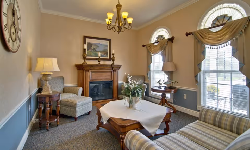 A cozy living room with a wooden fireplace mantel, two armchairs, a striped sofa, and a wooden coffee table with a white tablecloth and a flower arrangement. The room has two large arched windows with decorative curtains, a wall clock, a table lamp, and a chandelier hanging from the ceiling.