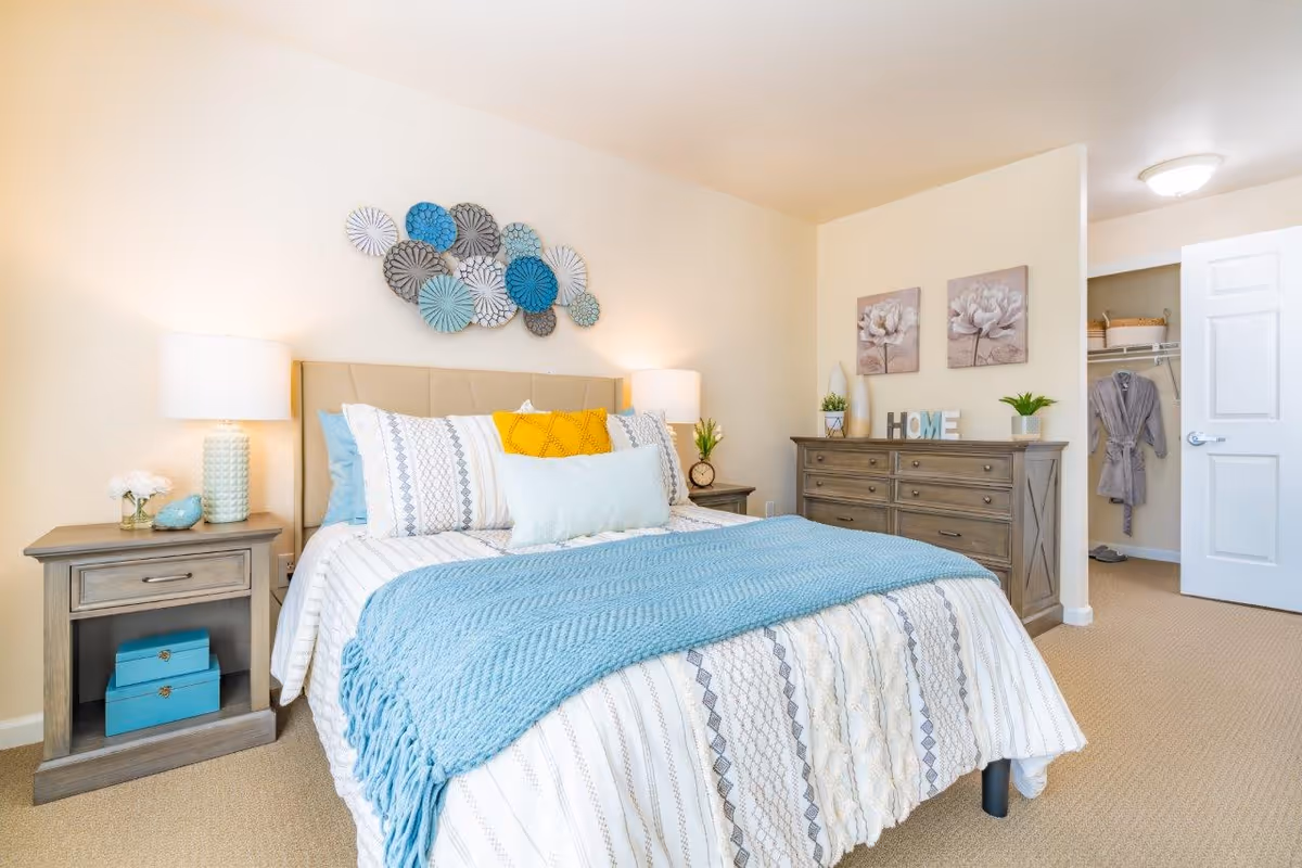 A bright and cozy bedroom featuring a neatly made bed with patterned bedding and a blue throw blanket. There are two bedside tables with lamps and decorative items. On the wall above the bed is a decorative arrangement of circular art pieces in shades of blue and gray. Across from the bed is a wooden dresser with plants, decorative letters spelling 'HOME', and floral artwork above it. An open door reveals a walk-in closet with a robe hanging inside.