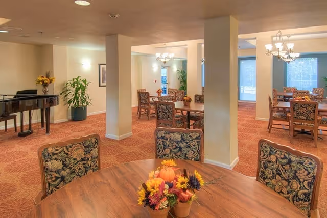 A spacious dining area in a retirement facility with round wooden tables and floral upholstered chairs. Each table has a small autumn-themed flower arrangement. The room has beige walls, carpeted floors with a red pattern, and several columns. There are chandeliers hanging from the ceiling and large windows letting in natural light. A black grand piano and some potted plants are visible in the background.