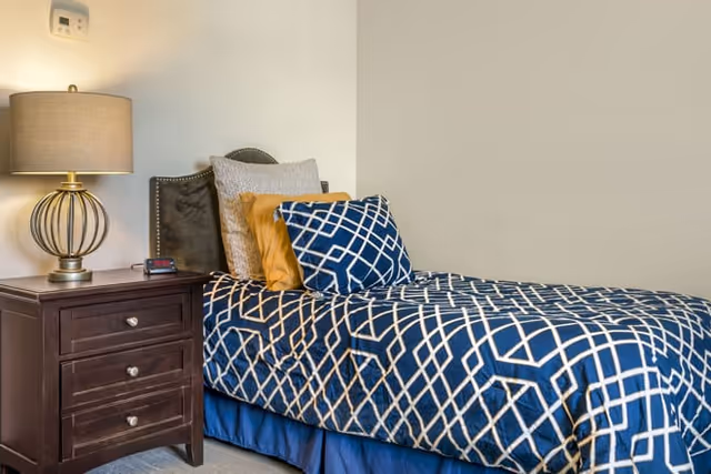 Single bed with blue geometric bedding and pillows beside a wooden nightstand with a lamp in a neutral bedroom.