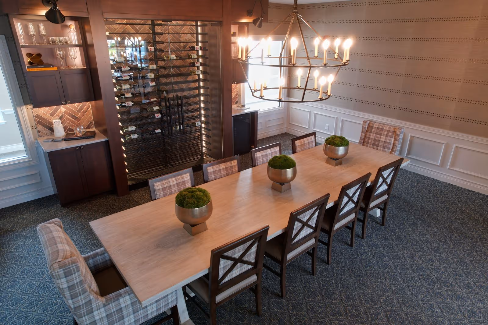 A dining room with a long wooden table surrounded by eight chairs, six with wooden frames and two with plaid upholstery. Three decorative bowls with green moss are placed on the table. A large chandelier with multiple candle-like lights hangs above the table. In the background, there is a wine rack filled with bottles and a cabinet with glassware. The room has patterned carpet, wainscoting on the walls, and a window letting in natural light.