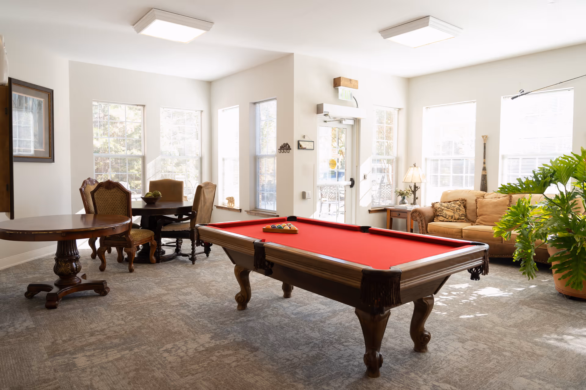 A bright and spacious common area with a red felt pool table in the center, a round wooden table with chairs near large windows, a beige sofa with patterned cushions, a side table with a lamp, and a large green potted plant. The room has white walls and carpeted flooring, with natural light streaming in through multiple windows.