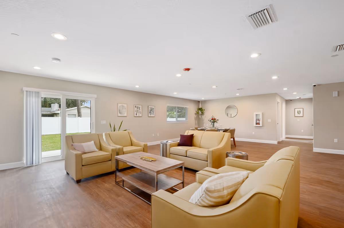 A spacious and well-lit living room area in a senior living facility featuring beige armchairs and sofas arranged around a wooden coffee table. The room has wood flooring, neutral-colored walls with framed artwork, a sliding glass door leading outside, and a dining table set with plates and glasses in the background.