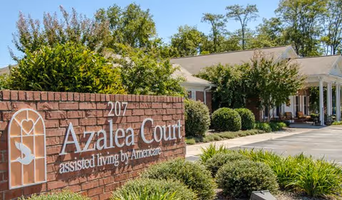 Exterior view of Azalea Court assisted living facility with a brick sign displaying the name and address '207 Azalea Court assisted living by Americare' surrounded by greenery and bushes, with the building and trees in the background under a clear sky.